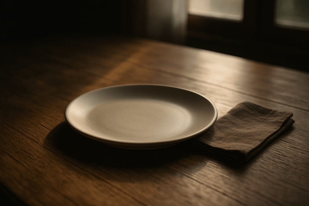 “Empty dinner plate on wooden table, soft morning light falling through window — symbol of remembrance and hunger.”
