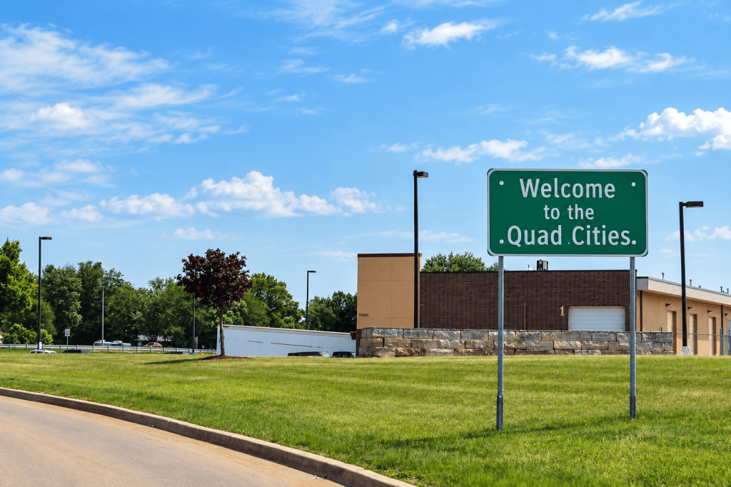 Welcome to the Quad Cities sign beside the road under a blue summer sky.